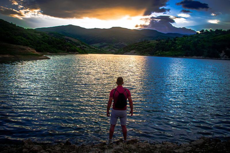 Rear view of man standing in lake against sky during sunset