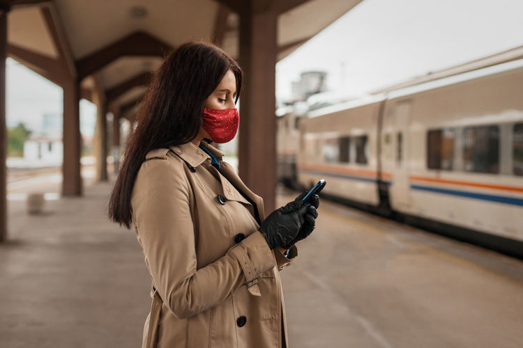 Side view of woman wearing mask using phone standing on railroad station platform