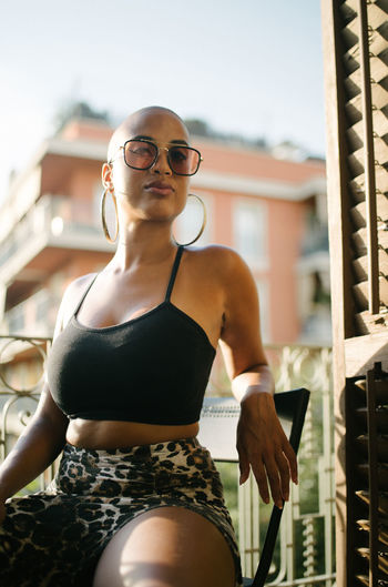 Low angle of black female with bald hairstyle and in trendy outfit sitting on terrace of outdoors cafe and relaxing while looking at camera
