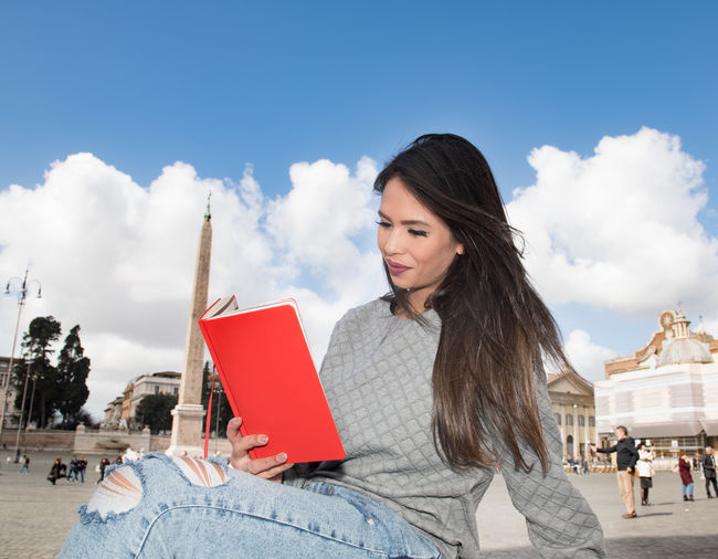Smiling young woman reading book while sitting against monument