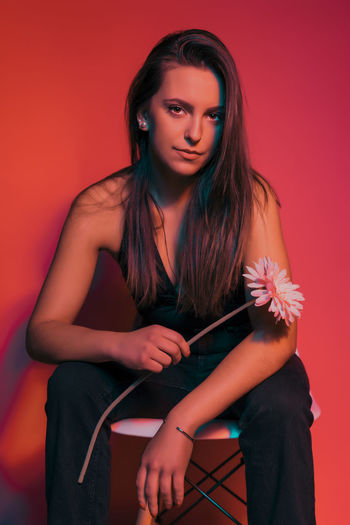 Portrait of young woman holding flower while sitting against red background