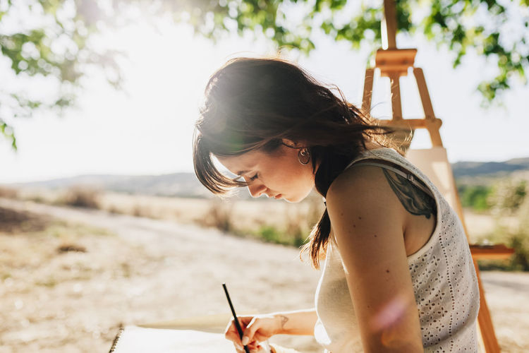 Young woman artist drawing in spiral notebook while sitting next to easel