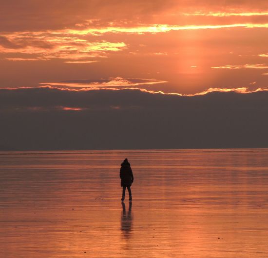 Silhouette man standing on frozen lake orange sky