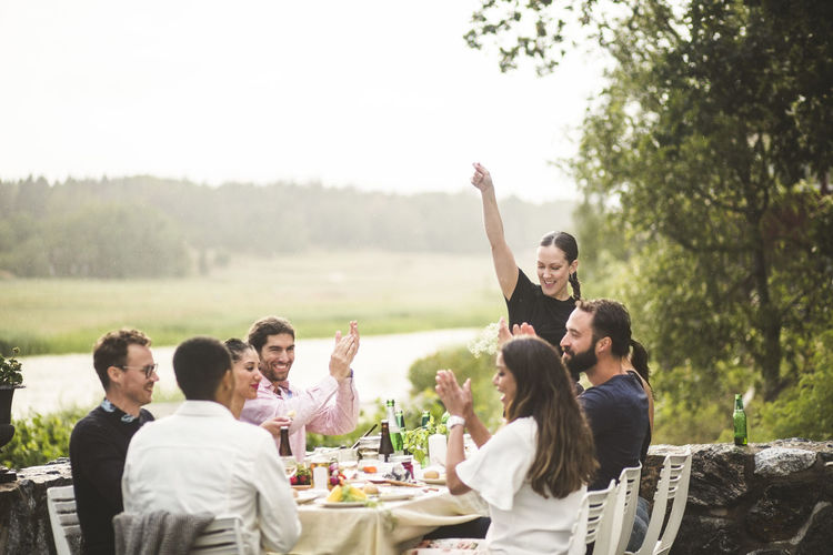 Mid adult woman standing with arm raised while friends applauding at table during dinner party in backyard