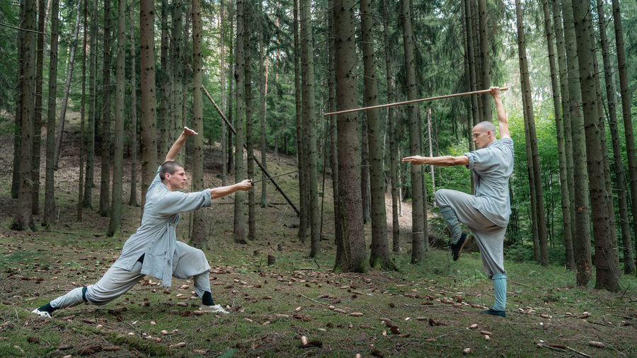 Full body men in gray clothes practicing kung fu with stick and sword during training in woods