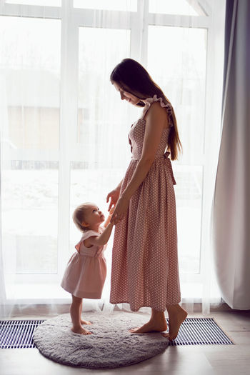 A young woman stands at a large window with a girl child in pink dresses at home barefoot