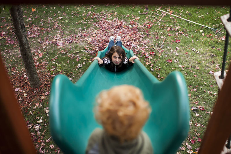 Girl slides down slide while looking up at her brother with pouty face