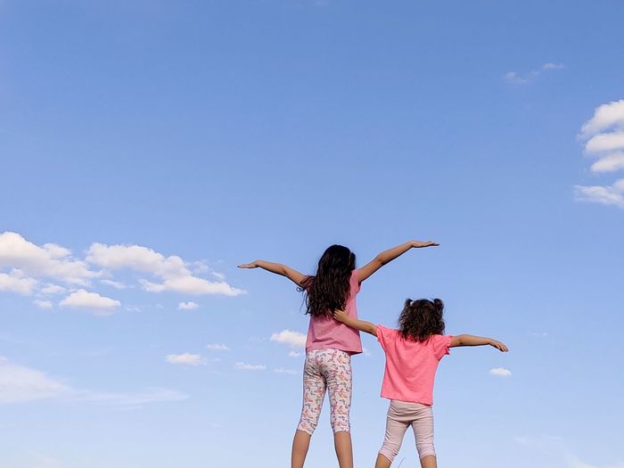 Low angle view of two girls with arms outstretched standing against blue sky