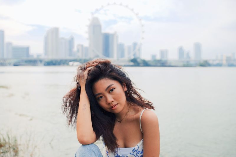 Portrait of young woman with hand in hair sitting against sea and urban skyline