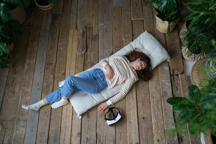 Relaxed woman resting after vr session, sleeping on wooden floor surrounded by tropical plants