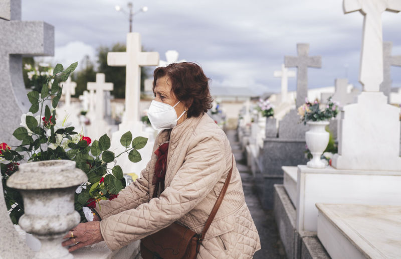Senior woman wearing protective face mask putting flowers on grave in cemetery