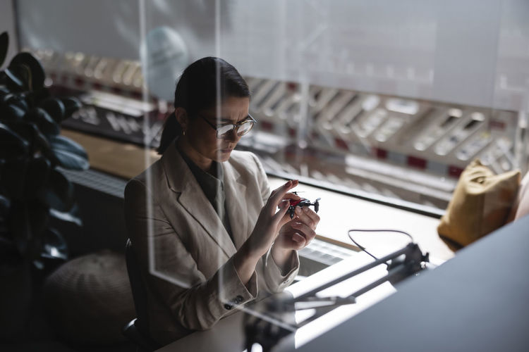 Businesswoman examining small drone at desk in office seen through glass