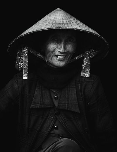 Portrait of senior woman wearing asian style conical hat against black background