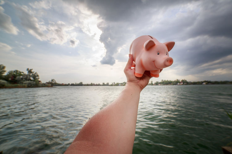 Cropped hand holding piggy bank over lake against cloudy sky