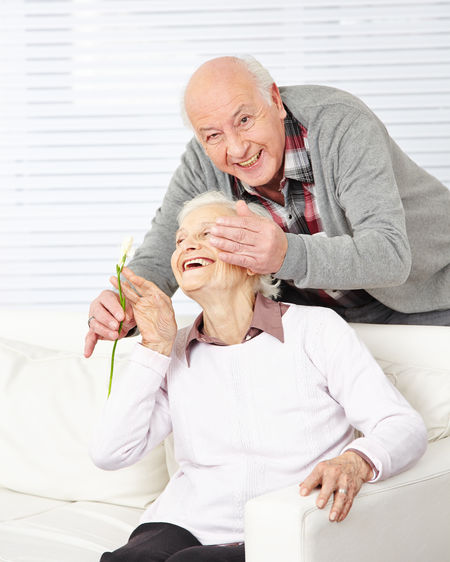 Portrait of smiling senior man holding flower embracing with woman while sitting on sofa at home