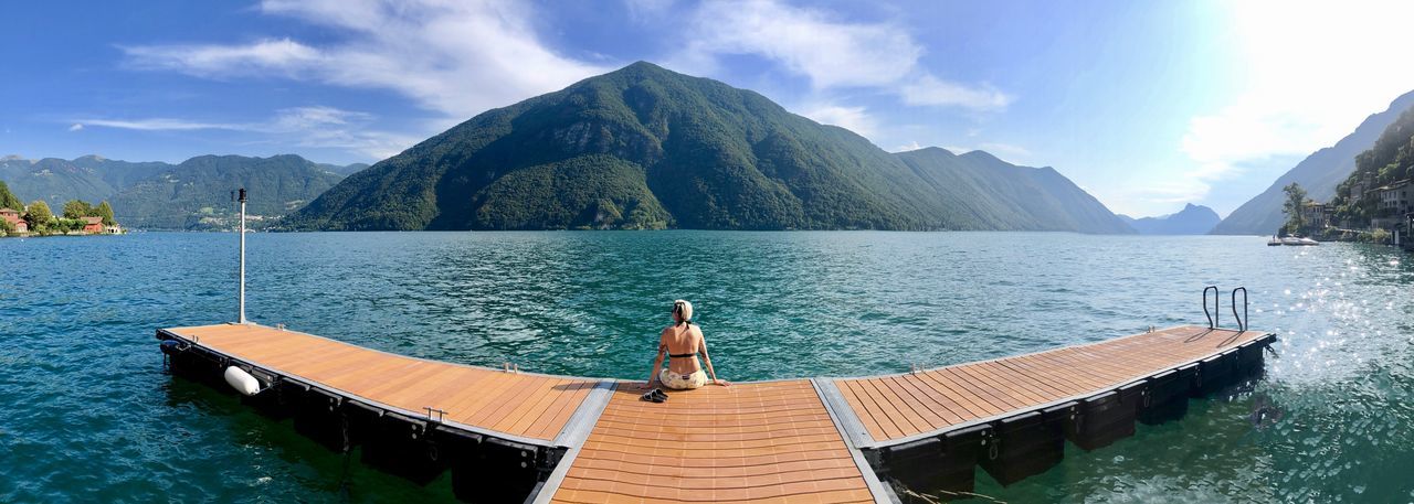 Rear view of woman sitting on pier over sea against mountains and sky