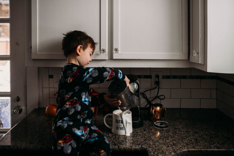Young boy sitting on kitchen couter spilling milk on coffee mug