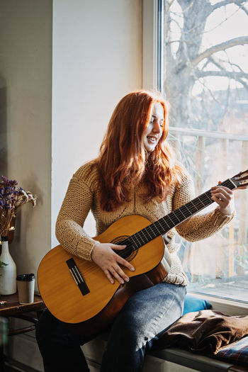 Unaltered candid portrait of young red haired woman in sweater playing acoustic guitar sitting by