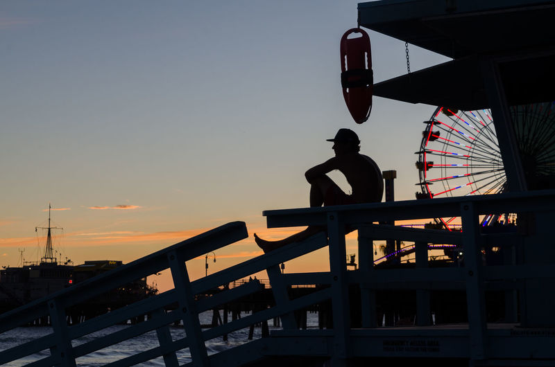 Silhouette man sitting at santa monica pier against sky during sunset