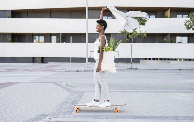 Businesswoman holding blazer while skateboarding with bag of vegetables on footpath