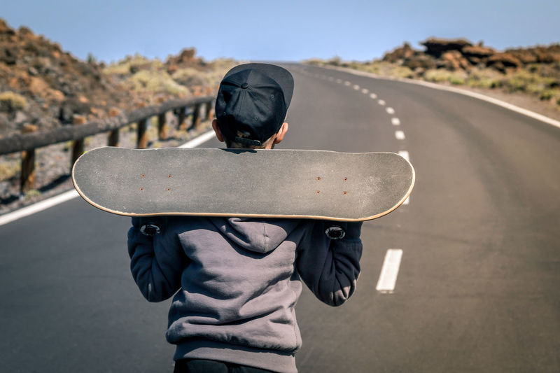 Rear view of boy standing with skateboard on road 