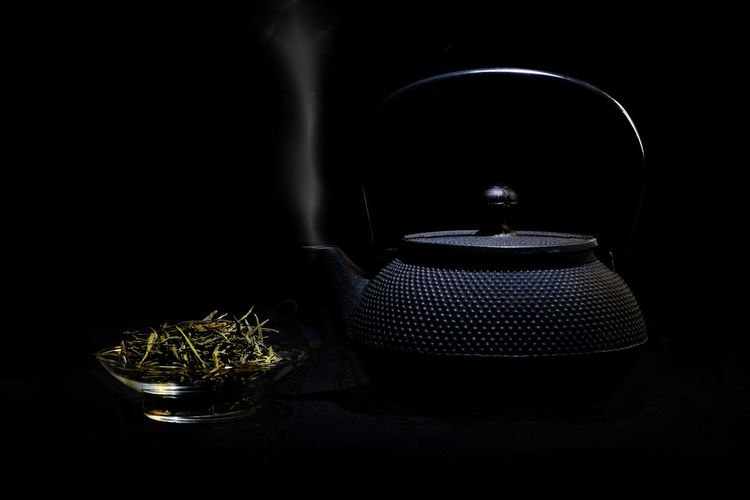 Green tea leaves in bowl by teapot emitting smoke against black background