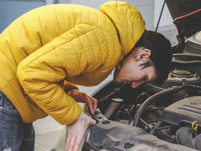 A  caucasian guy in a yellow jacket inspects an internal breakdown with an open car hood of his car