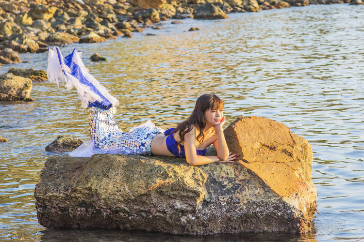 Smiling woman in mermaid costume relaxing on rock in lake