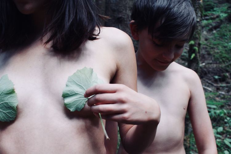 Midsection of shirtless woman covering breast with leaves by boy in forest
