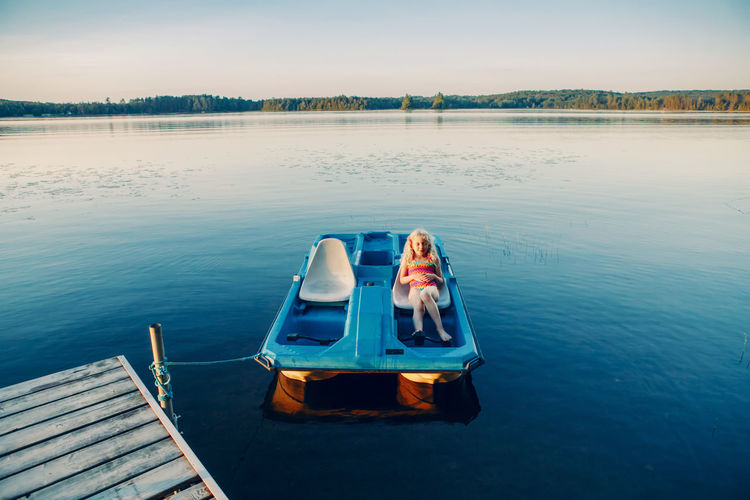 Child girl sitting relaxing in old  pedal boat tied to wooden dock pier. beautiful idyllic landscape 
