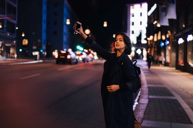 Young woman with phone hailing for taxi in city at night