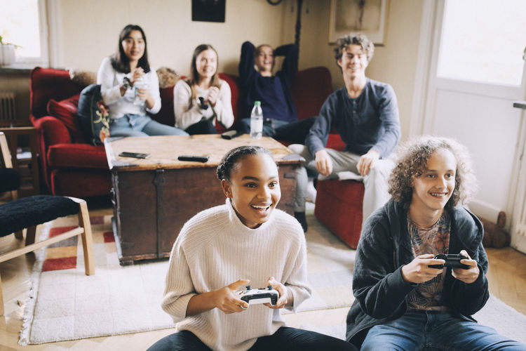 Happy teenagers playing video game while friends sitting on sofa in living room