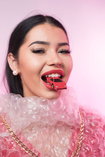 Portrait of young woman wearing bubble wrap with toy car in mouth against pink background