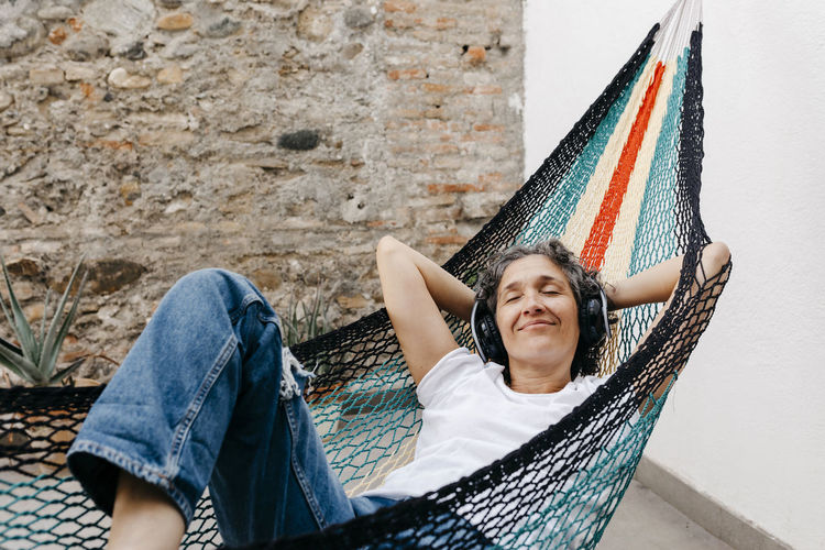Woman listening music through headphones while relaxing on hammock against wall at back yard