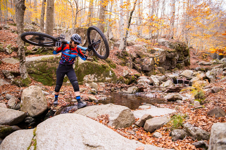 Full body woman in helmet and sportswear carrying mountain bike on shoulders while crossing rocky creek in autumn forest
