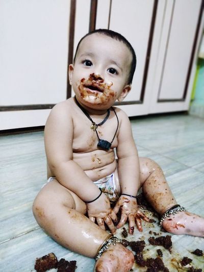 Cute baby girl eating cake with messy face while sitting on floor at home