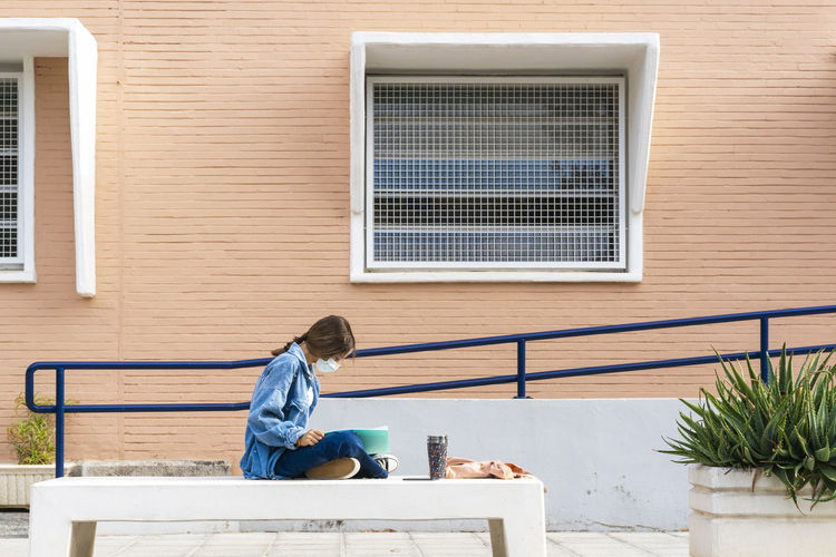 Teenage girl wearing protective face mask reading book while sitting on concrete mask