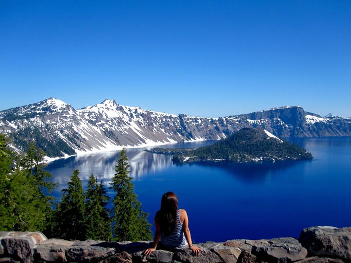 Woman sitting at lakeshore against snowcapped mountains