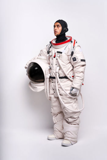 Full body of colombian male astronaut in spacesuit with helmet in hand standing in studio against white background and looking away