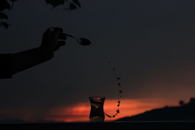 Cropped hand pouring drink in glass against cloudy sky during sunset