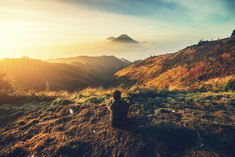 Rear view of man sitting on mountain against sky during sunset