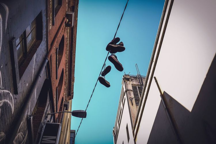 Low angle view of shoes hanging on cable by building against clear blue sky