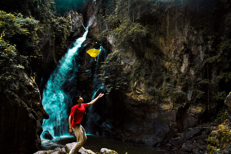 Man throwing hat in mid-air while standing on rock against waterfall