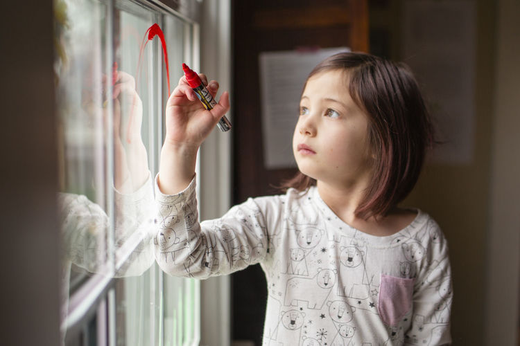A serious little girl draws on a window with a bright red marker