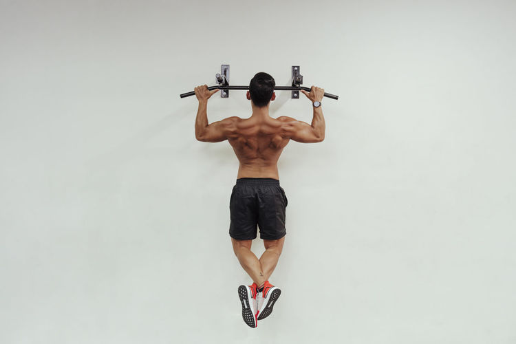 Mid adult sportsman doing chin-ups with exercise equipment on white wall