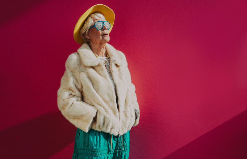 Senior woman wearing hat and sunglasses standing against pink background