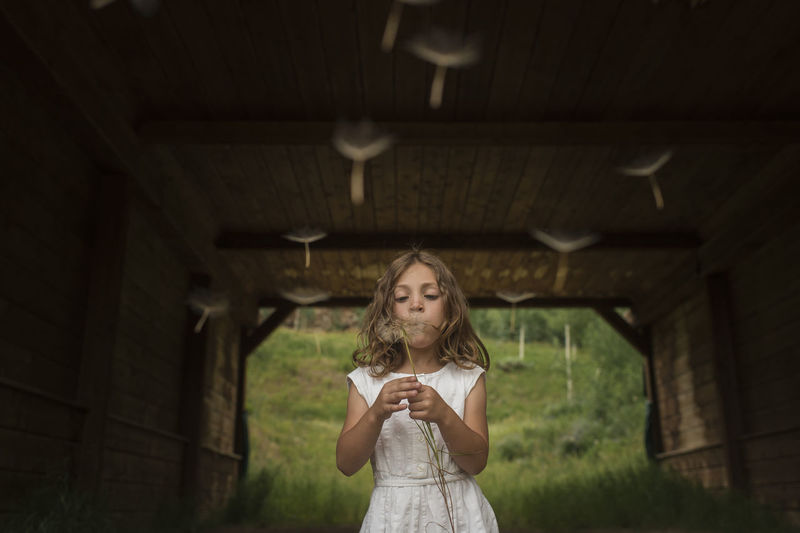 Girl blowing dandelion seeds while standing under shed