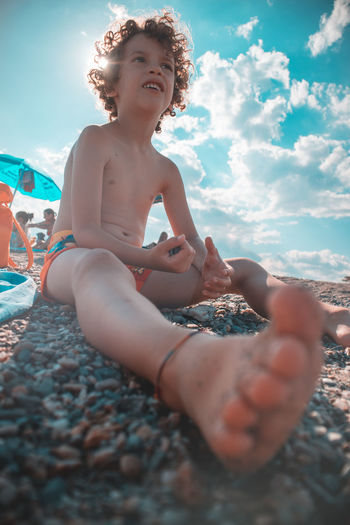 Full length of shirtless boy sitting on pebbles at beach against sky