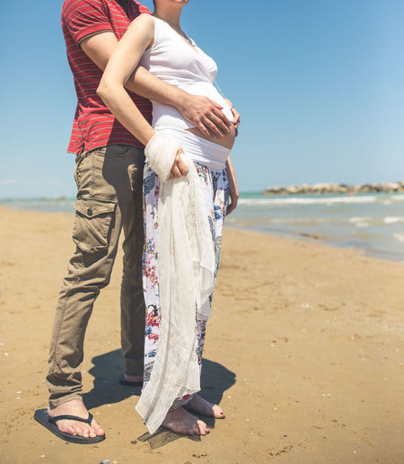 Low section of man holding pregnant woman belly at beach