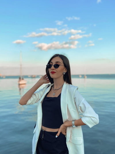 Young woman wearing sunglasses standing at sea against sky